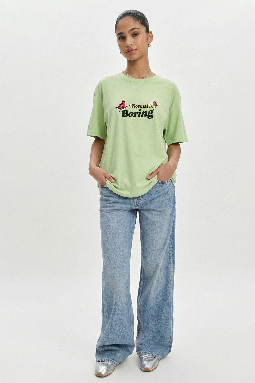 Person wearing a light green t-shirt with text and blue jeans on a white background.pastel green oversized t-shirt, normal is boring graphic tee, butterfly back print t-shirt, floral aesthetic oversized tee, dopamine dressing outfit, gen z streetwear india, oversized cotton graphic t-shirt women, aesthetic back print tee, casual summer oversized t-shirt, trendy statement t-shirt