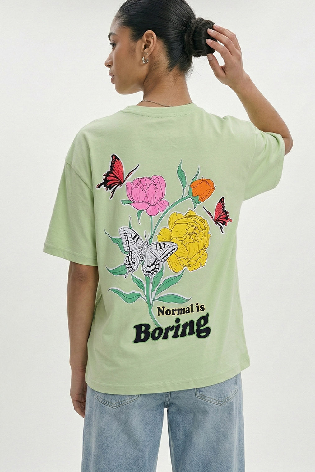 Person wearing a light green t-shirt with floral and butterfly design and 'Normal is Boring' text.pastel green oversized t-shirt, normal is boring graphic tee, butterfly back print t-shirt, floral aesthetic oversized tee, dopamine dressing outfit, gen z streetwear india, oversized cotton graphic t-shirt women, aesthetic back print tee, casual summer oversized t-shirt, trendy statement t-shirt.