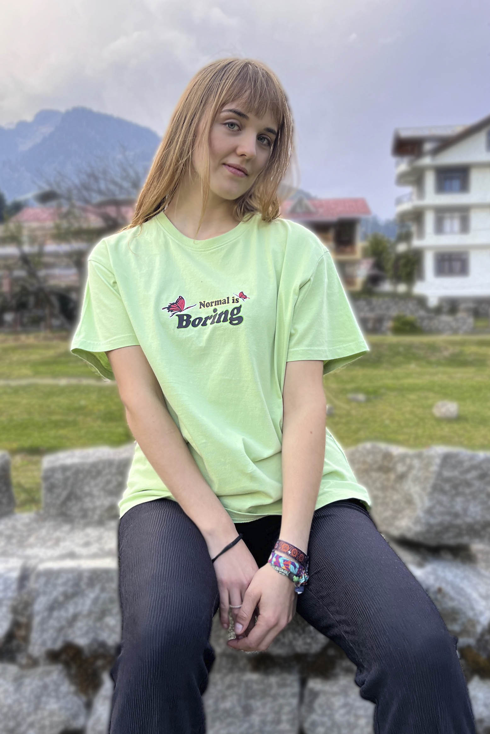 A woman sitting on a stone bench wearing a pastel green oversized t-shirt with a phrase printed on the front and a lit flower and butterfly graphic on the back.
