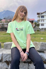 A woman sitting on a stone bench wearing a pastel green oversized t-shirt with a phrase printed on the front and a lit flower and butterfly graphic on the back.