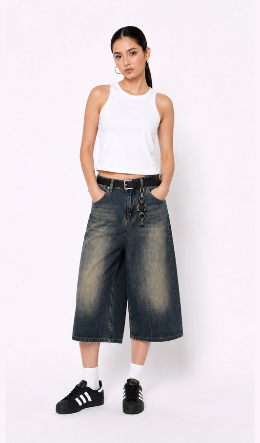 Woman wearing a white sleeveless top, dark blue denim culottes(jorts), and black sneakers on a white background.white tank top, white tank top women, women's white tank top, sleeveless white top, basic white tank top, white camisole top, white summer top, women's casual top, white fashion top, dark blue denim culottes, denim culottes women, denim jorts women, women's jorts outfit, wide leg denim shorts, knee length denim shorts, blue denim jorts, denim bermuda shorts women, culotte jeans.