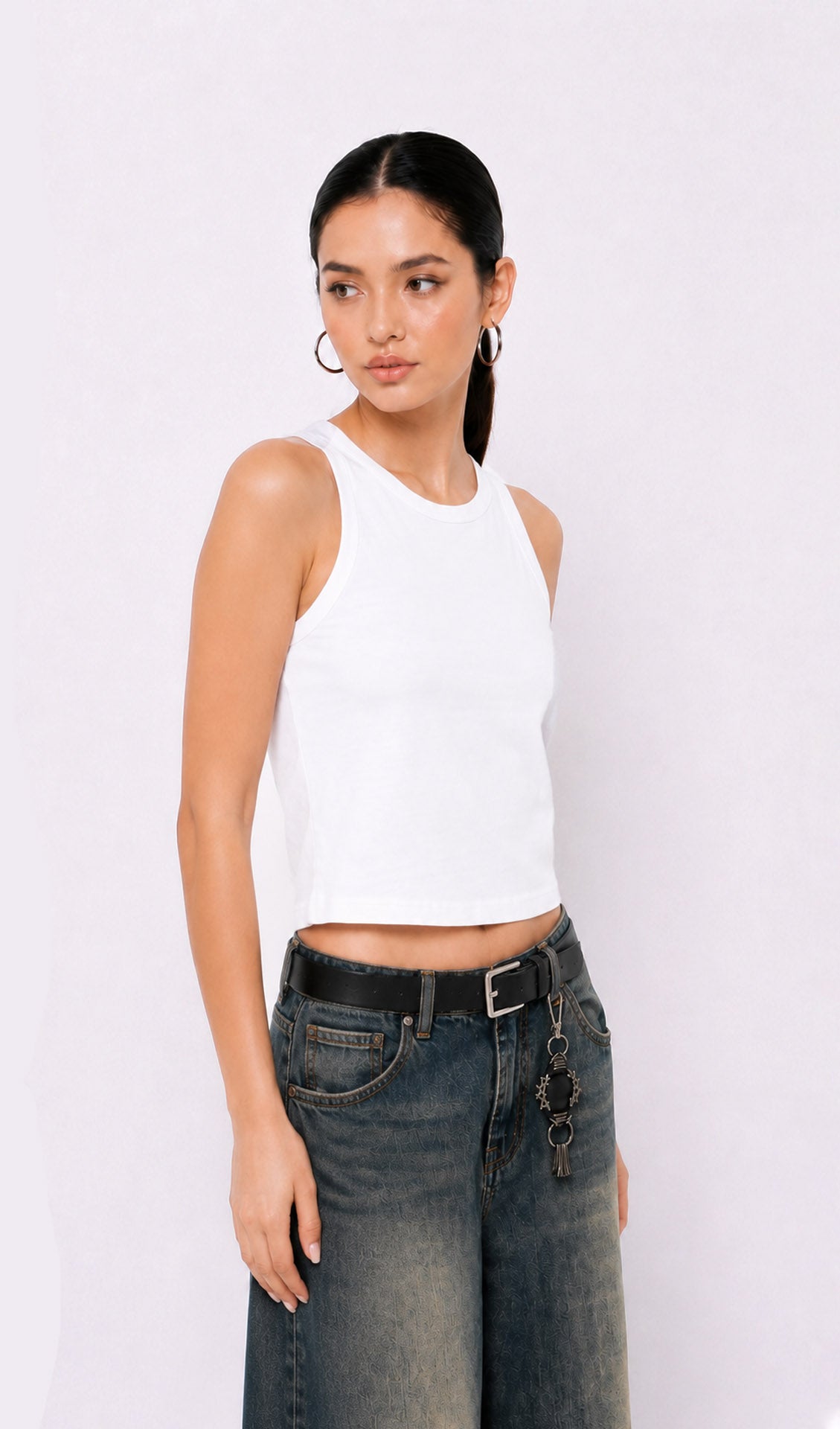 Woman wearing a white sleeveless top and dark jeans on a plain background.white tank top, white tank top women, women's white tank top, sleeveless white top, basic white tank top, white camisole top, white summer top, women's casual top, white fashion top, dark blue denim culottes, denim culottes women, denim jorts women, women's jorts outfit, wide leg denim shorts, knee length denim shorts, blue denim jorts, denim bermuda shorts women, culotte jeans.
