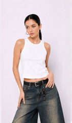 Woman wearing a white tank top and high-waisted jeans against a light purple background.white tank top, white tank top women, women's white tank top, sleeveless white top, basic white tank top, white camisole top, white summer top, women's casual top, white fashion top, dark blue denim culottes, denim culottes women, denim jorts women, women's jorts outfit, wide leg denim shorts, knee length denim shorts, blue denim jorts, denim bermuda shorts women, culotte jeans.