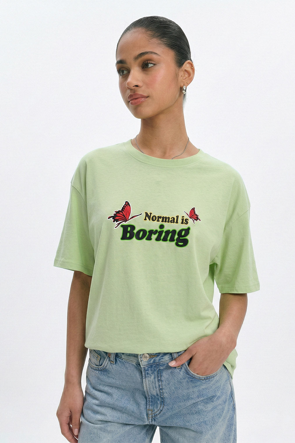 Person wearing a light green t-shirt with 'Normal is Boring' text and butterfly graphics on a white background. pastel green oversized t-shirt, normal is boring graphic tee, butterfly back print t-shirt, floral aesthetic oversized tee, dopamine dressing outfit, gen z streetwear india, oversized cotton graphic t-shirt women, aesthetic back print tee, casual summer oversized t-shirt, trendy statement t-shirt.