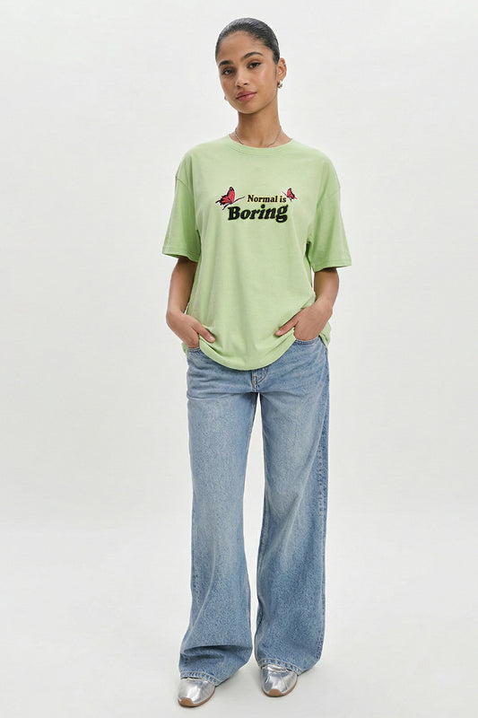 Person wearing a light green t-shirt with text and blue jeans on a white background.pastel green oversized t-shirt, normal is boring graphic tee, butterfly back print t-shirt, floral aesthetic oversized tee, dopamine dressing outfit, gen z streetwear india, oversized cotton graphic t-shirt women, aesthetic back print tee, casual summer oversized t-shirt, trendy statement t-shirt 