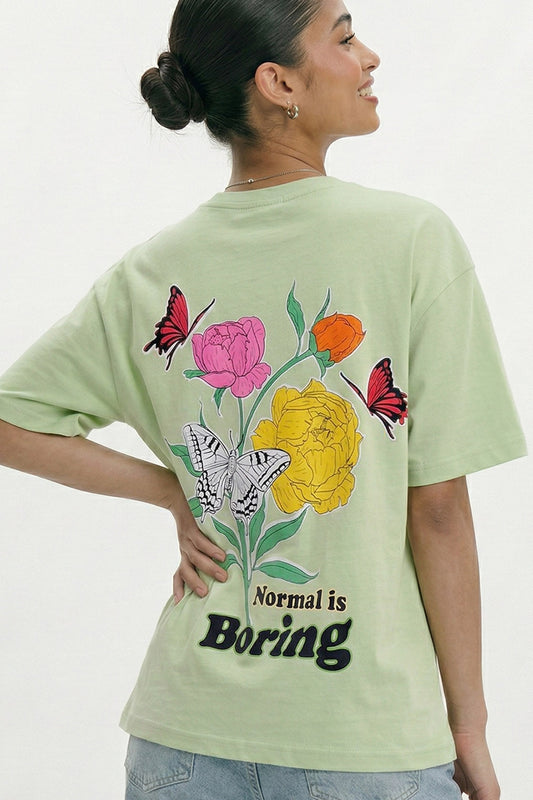Person wearing a light green t-shirt with floral and butterfly design and 'Normal is Boring' text.pastel green oversized t-shirt, normal is boring graphic tee, butterfly back print t-shirt, floral aesthetic oversized tee, dopamine dressing outfit, gen z streetwear india, oversized cotton graphic t-shirt women, aesthetic back print tee, casual summer oversized t-shirt, trendy statement t-shirt