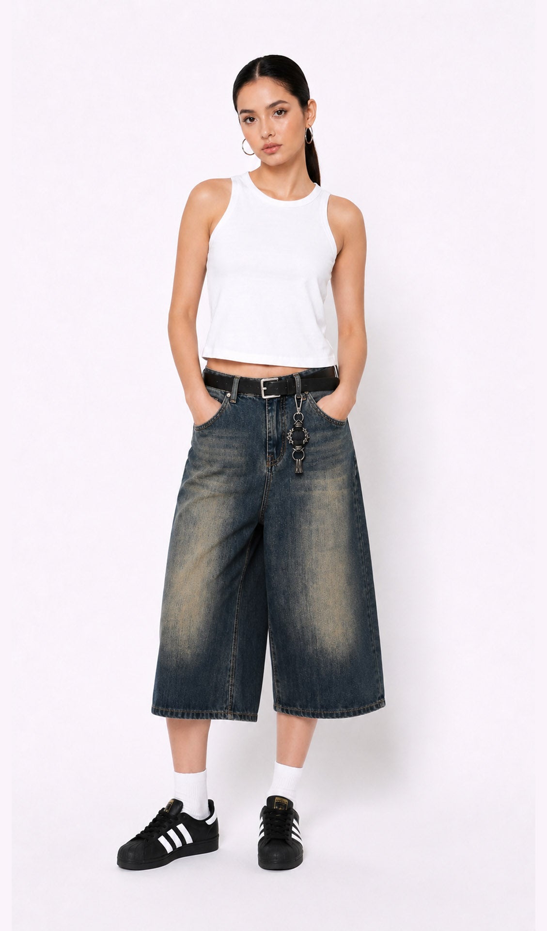 Woman wearing a white sleeveless top, dark blue denim culottes(jorts), and black sneakers on a white background.white tank top, white tank top women, women's white tank top, sleeveless white top, basic white tank top, white camisole top, white summer top, women's casual top, white fashion top, dark blue denim culottes, denim culottes women, denim jorts women, women's jorts outfit, wide leg denim shorts, knee length denim shorts, blue denim jorts, denim bermuda shorts women, culotte jeans.