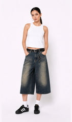 Woman wearing a white sleeveless top, dark blue denim culottes(jorts), and black sneakers on a white background.white tank top, white tank top women, women's white tank top, sleeveless white top, basic white tank top, white camisole top, white summer top, women's casual top, white fashion top, dark blue denim culottes, denim culottes women, denim jorts women, women's jorts outfit, wide leg denim shorts, knee length denim shorts, blue denim jorts, denim bermuda shorts women, culotte jeans.